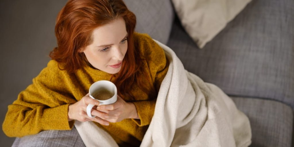 Relaxed young redhead woman enjoying a tea break sitting wrapped in a warm blanket on a comfortable couch 